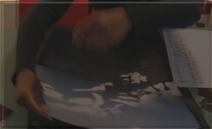 Chandra Brooks lifts a hand-cut paper light cutting during the making process, revealing intricate shadows cast onto the work surface.
